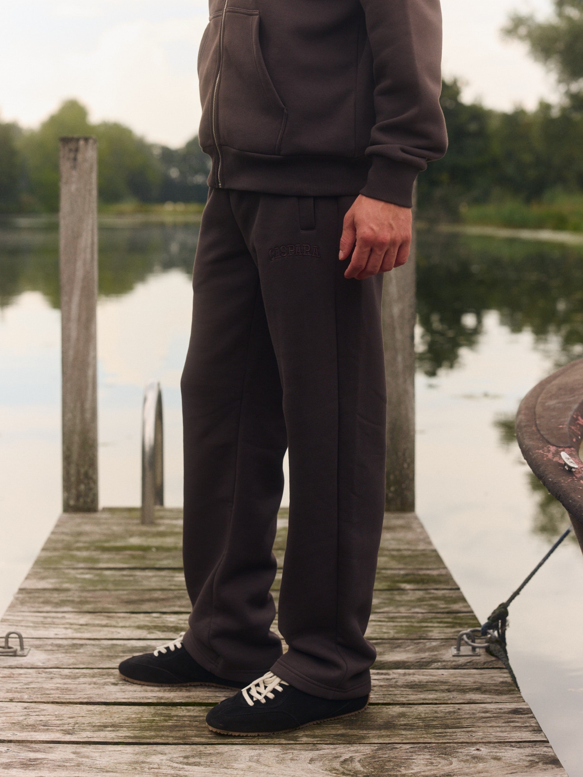 Man wearing espresso straight-leg sweatpants and matching zip hoodie standing on a wooden dock.